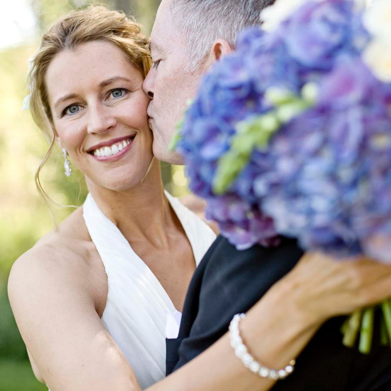 wedding photography of the groom kissing the bridges cheek outside while she smiles holding a purple bouquet of hydrangeas