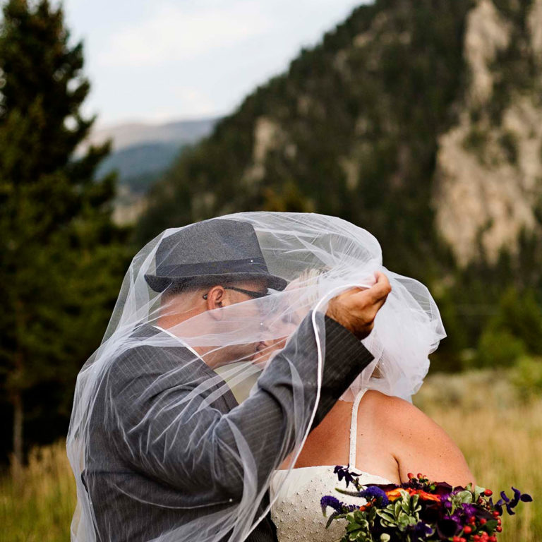wedding photography of the bride and groom kissing while the groom uses the bride's veil for privacy outside in the field