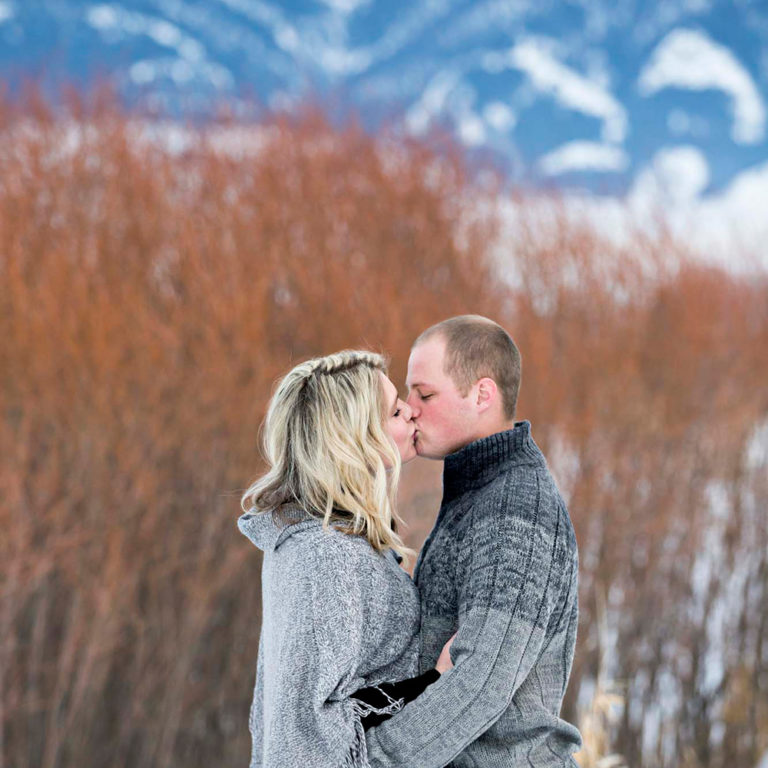 engagement photography of a couple bundled up outside in a snowy valley kissing in front of a mountain